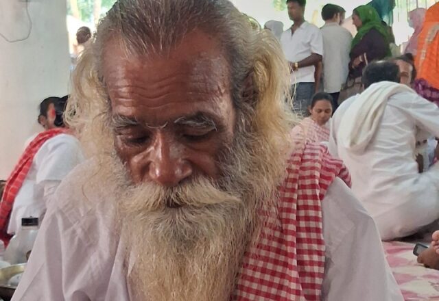 Baul elder with a braided beard and red checked gamchha at Lalon Akhra