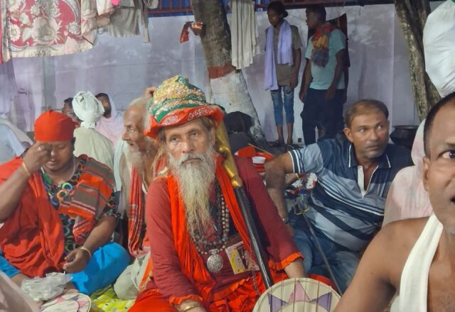 Baul fakir in saffron robes and a decorated turban among musicians at the Lalon Mela